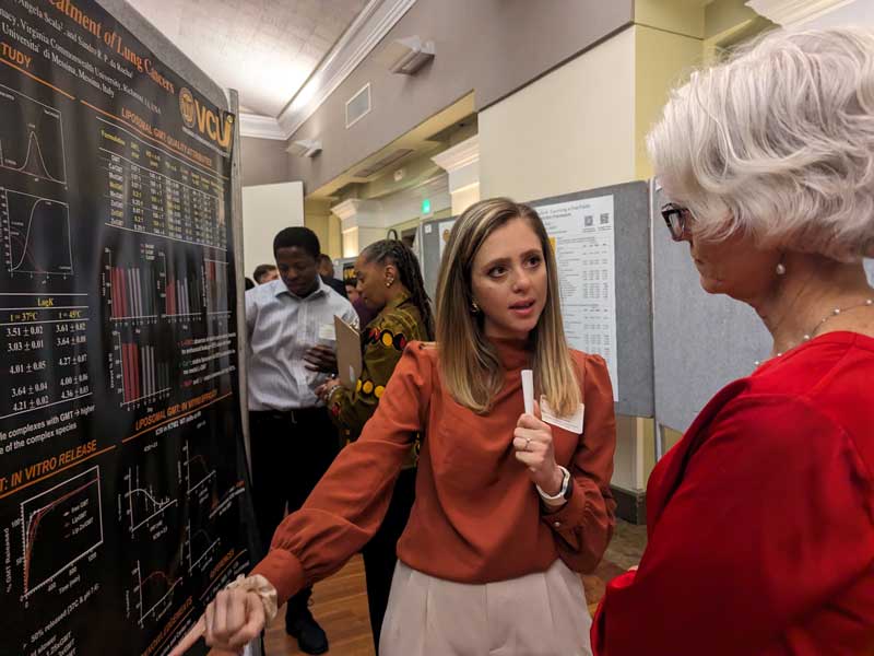 a student presenter points to a research poster while speaking to a professor
