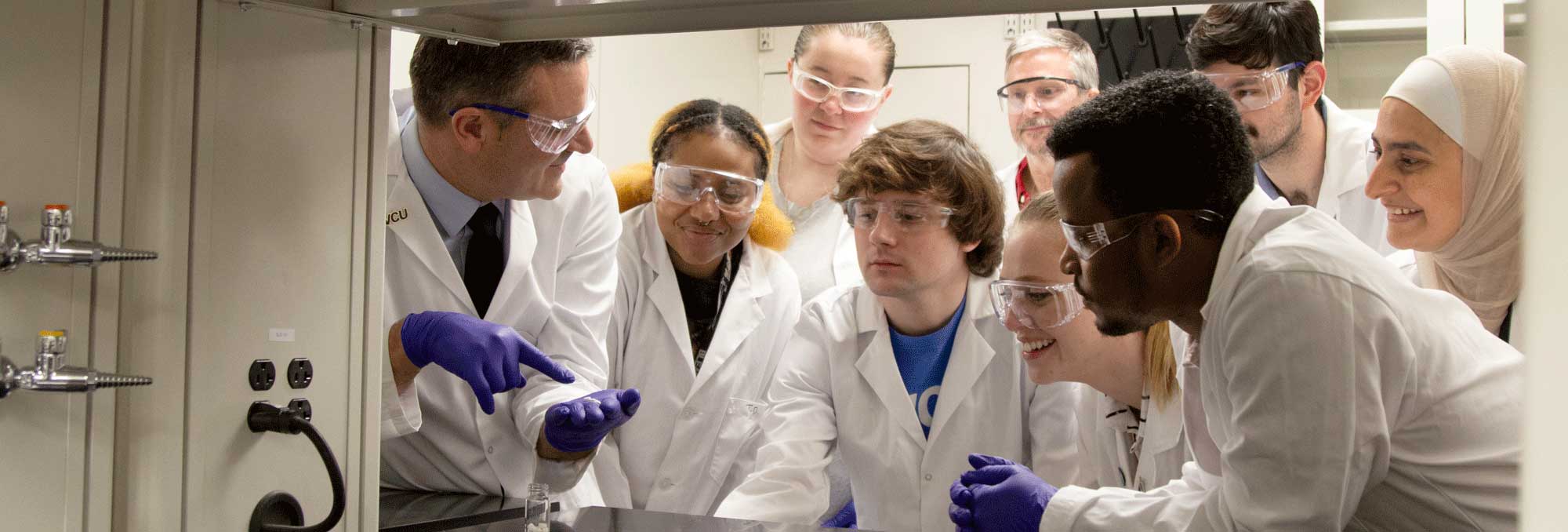 A professor and students in lab coats, gloves and protective gear gather in a lab to look at a sample of a medication.