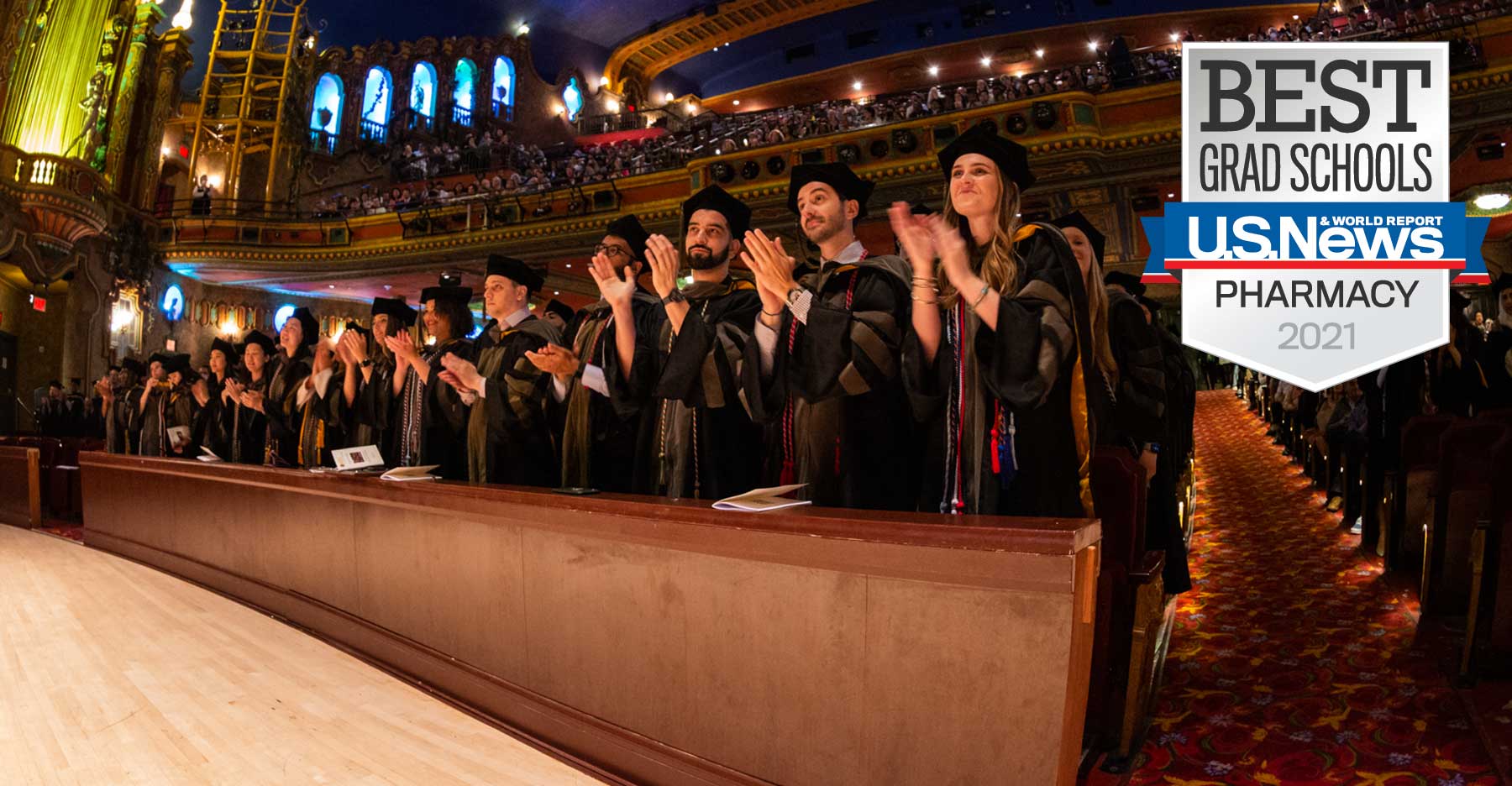 pharmacy student graduates standing and clapping at their graduation ceremony alongside a u.s. news and world reports banner ranking v.c.u. school of pharmacy as a best grad school for 2021