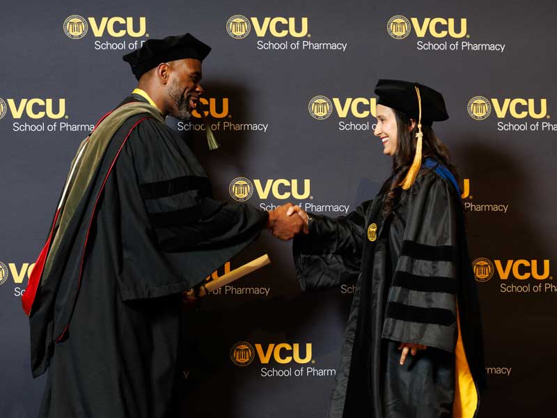 Graduating Ph.D. student Sagun Poudel in graduation regalia shakes hands with Dean Ogbonna in front of a VCU School of Pharmacy backdrop at the VCU School of Pharmacy Class of 2026 Hooding & Diploma Ceremony.