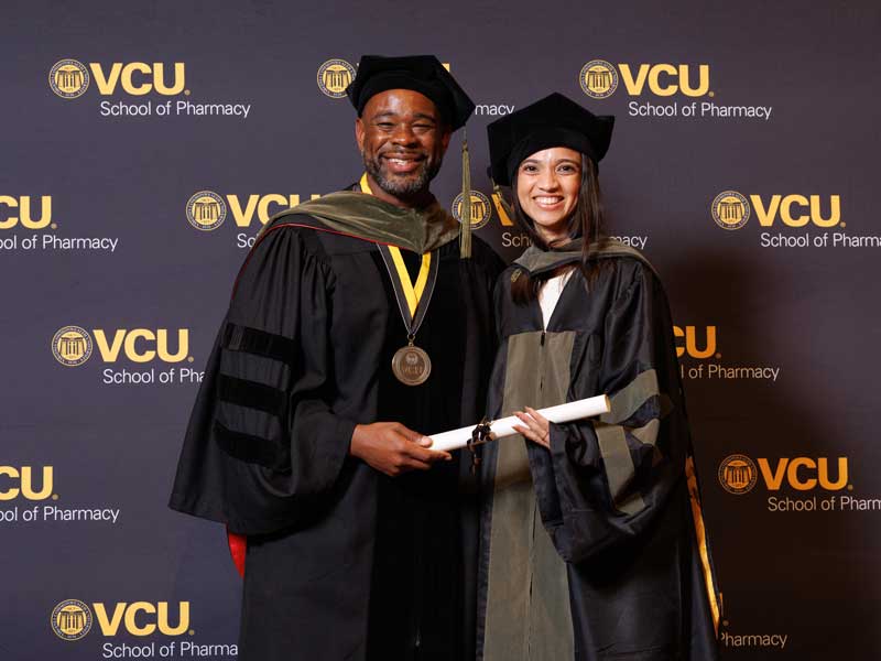 Basima Zaman in graduation regalia holds her diploma standing next to Dean Ogbonna in front of a VCU School of Pharmacy backdrop at the VCU School of Pharmacy Class of 2026 Hooding & Diploma Ceremony.