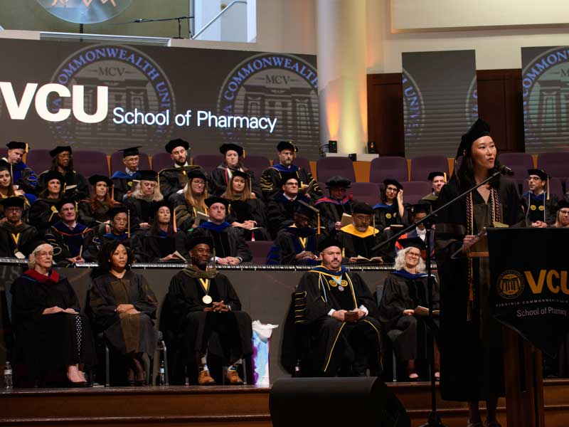 Pin-Hsuan Wu in graduation regalia addresses the audience at the VCU School of Pharmacy Class of 2026 Hooding & Diploma Ceremony in front of rows of faculty proudly looking on from behind her onstage.