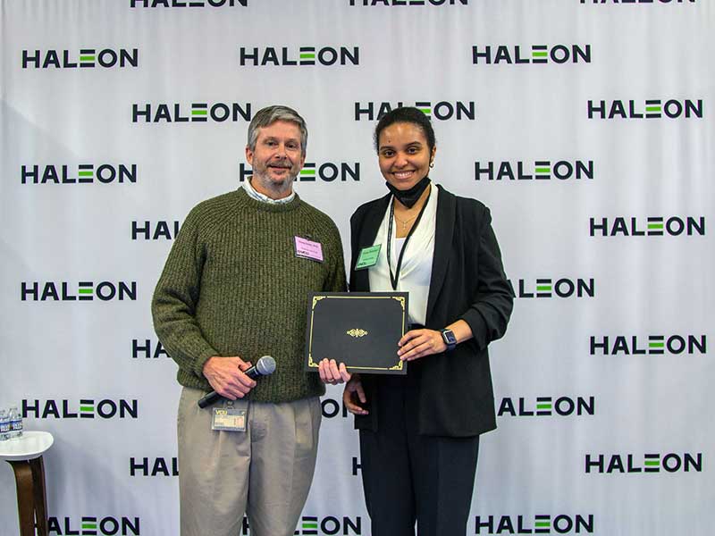 A student holding an award stands next to the department chair of Pharmaceutics in front of a backdrop that has the Haleon logo