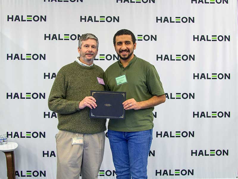 A student holding an award stands next to the department chair of Pharmaceutics in front of a backdrop that has the Haleon logo