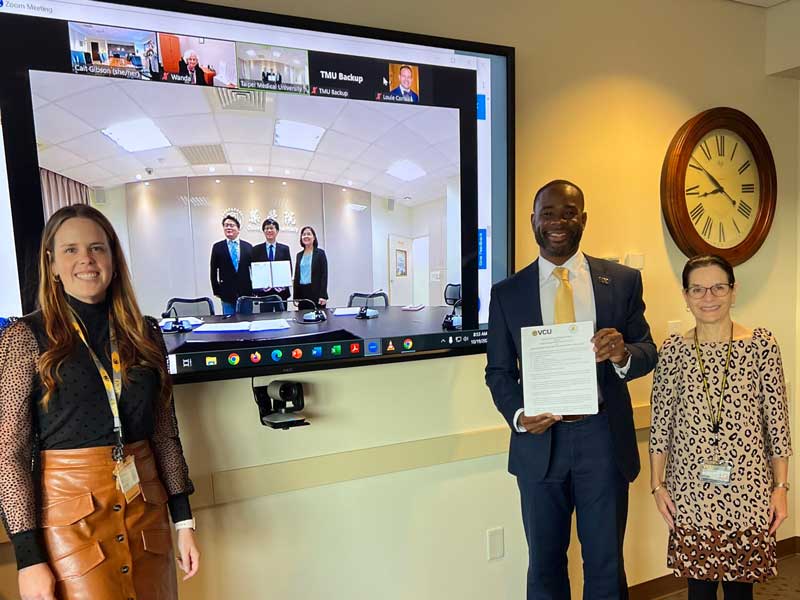 three representatives from the v.c.u. school of pharmacy hold up a signed agreement while representatives from taipei medical university hold up a signed agreement on a video feed displayed on a large screen