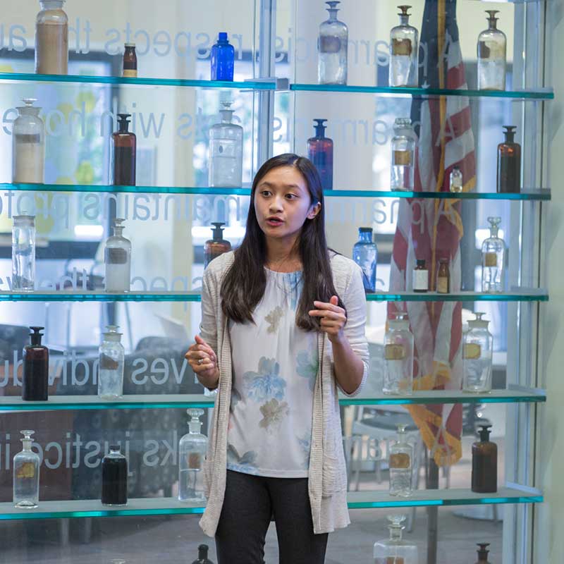 A student standing in front of V C U Pharmacy Smith building glass wall presenting.