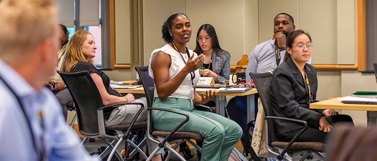 Several adults seated around tables in a classroom or conference room, participating in a group discussion. A woman in the center, wearing a white top and green pants, is speaking and gesturing with her hands. Other participants are listening, writing notes, or observing the conversation.