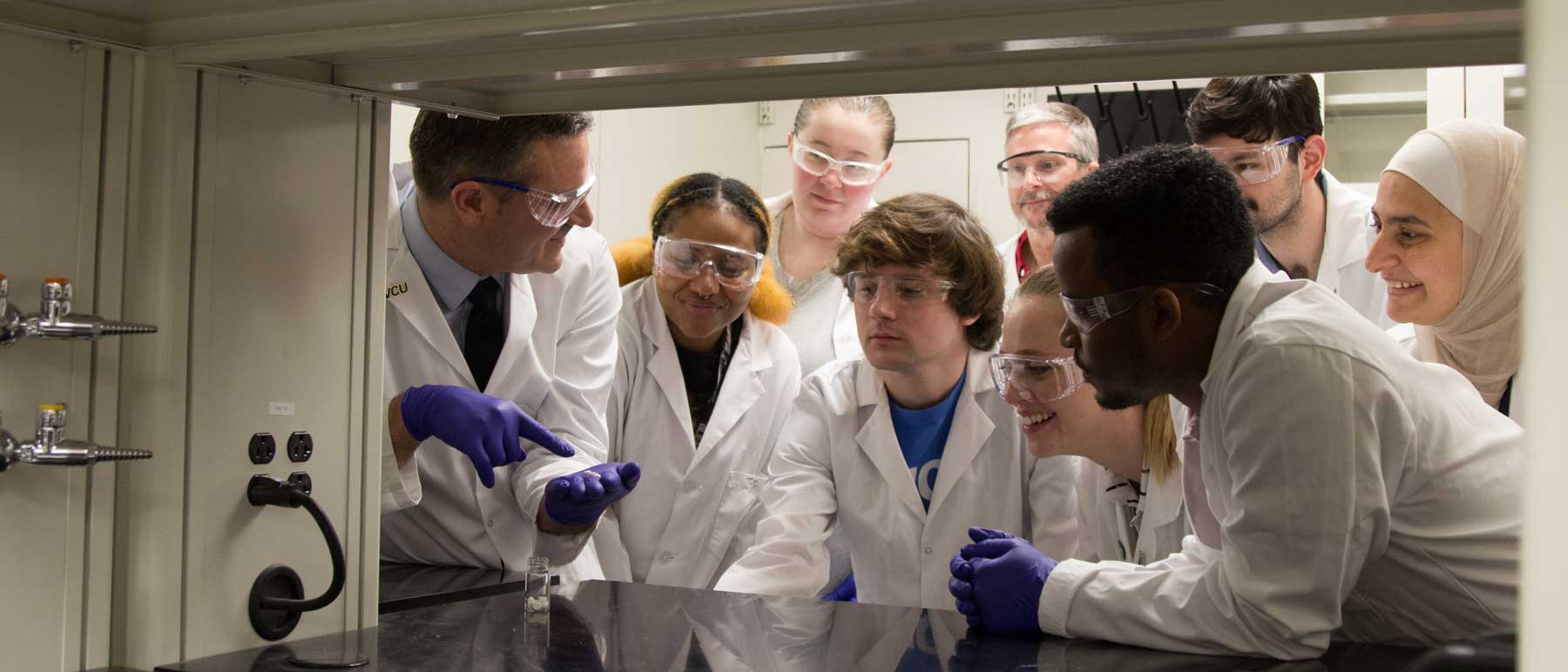 a professor points to a test tube in a lab while eager students observe and take notes