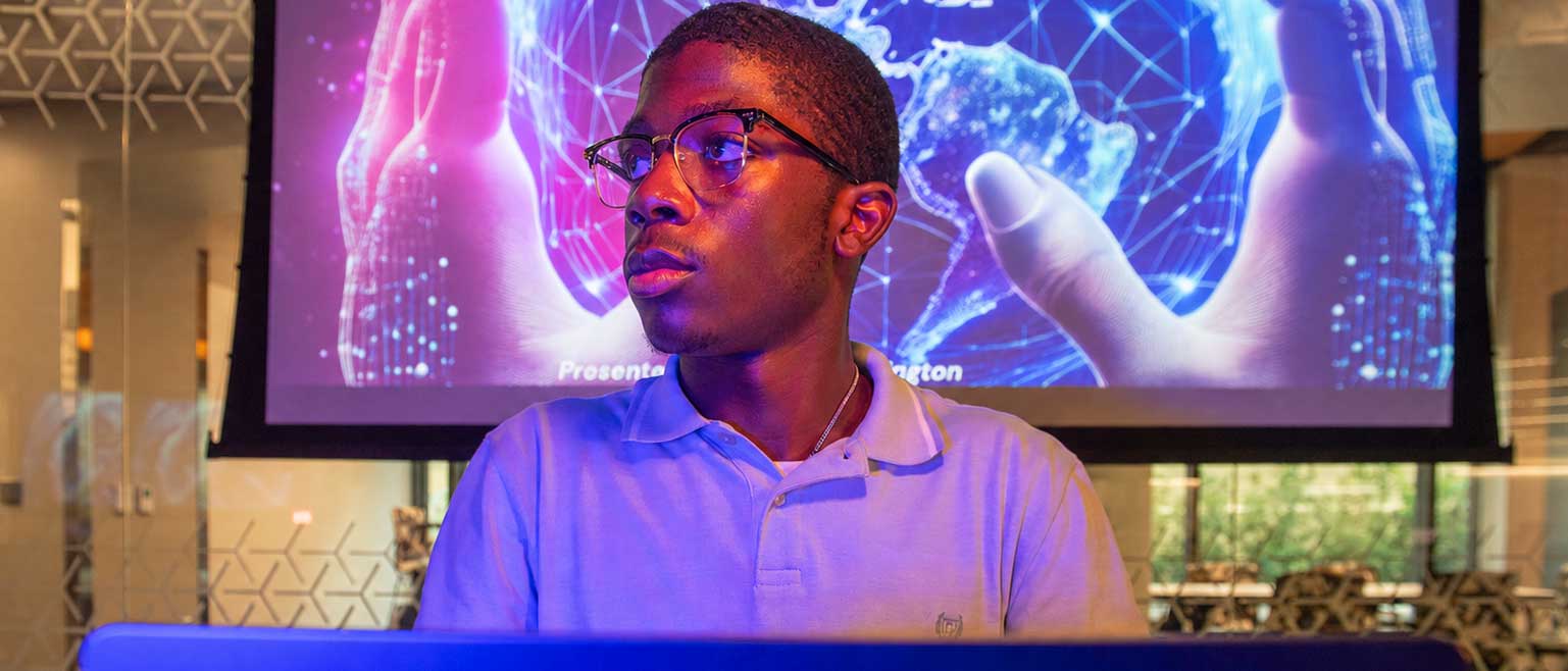 A person sits behind a laptop, looking to the side as a large digital display shows two hands surrounding a networked globe.
