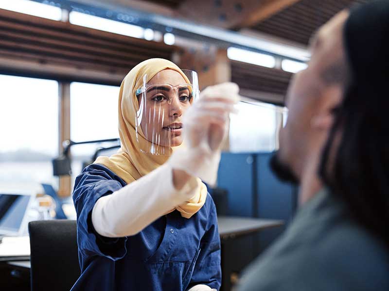 A pharmacist in a hijab, scrubs and gloves swabs a patient for strep throat.