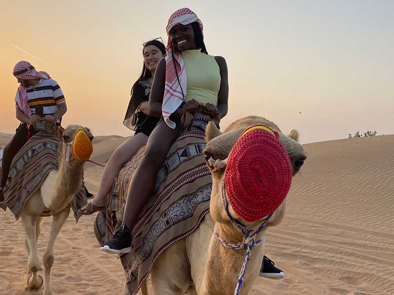 Two students in a sandy desert ride a camel at sunset