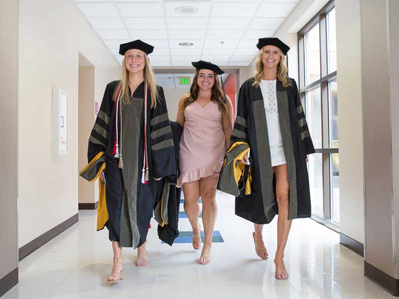 Three students walk through a hallway wearing graduation cap and gown.