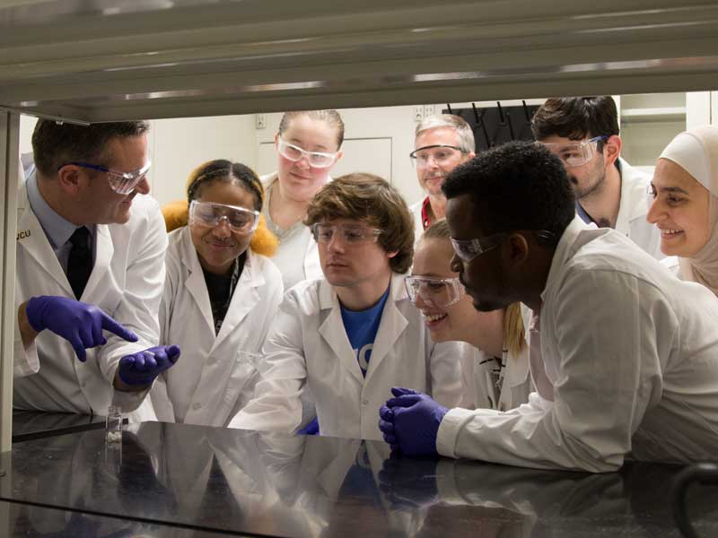 a professor points to a test tube in a lab while eager students observe and take notes