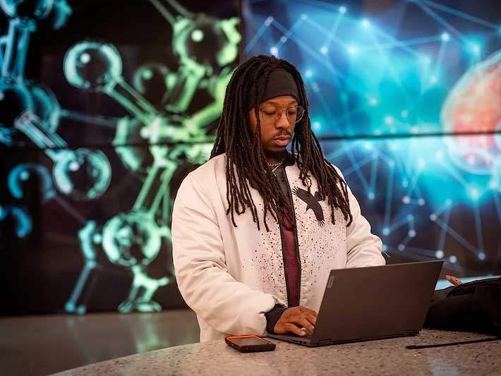 a student working on a laptop in front of a brightly colored wall featuring large displays of science images such as d.n.a. strands and molecules