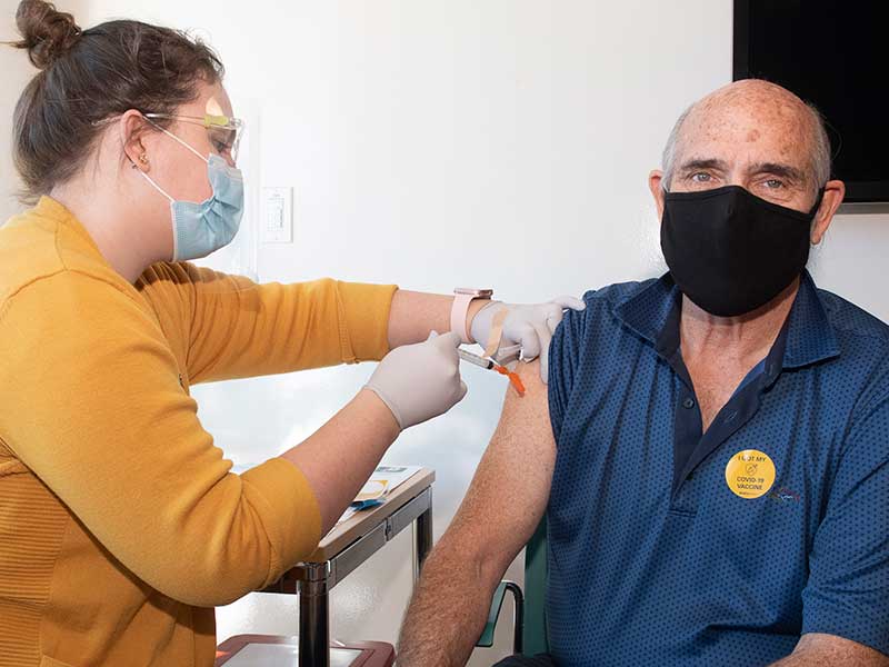 A VCU student with a gold sweater, a mask and her hair in a bun administers the COVID-19 vaccination to an older adult man at the VCU Health Hub at 25th in 2021