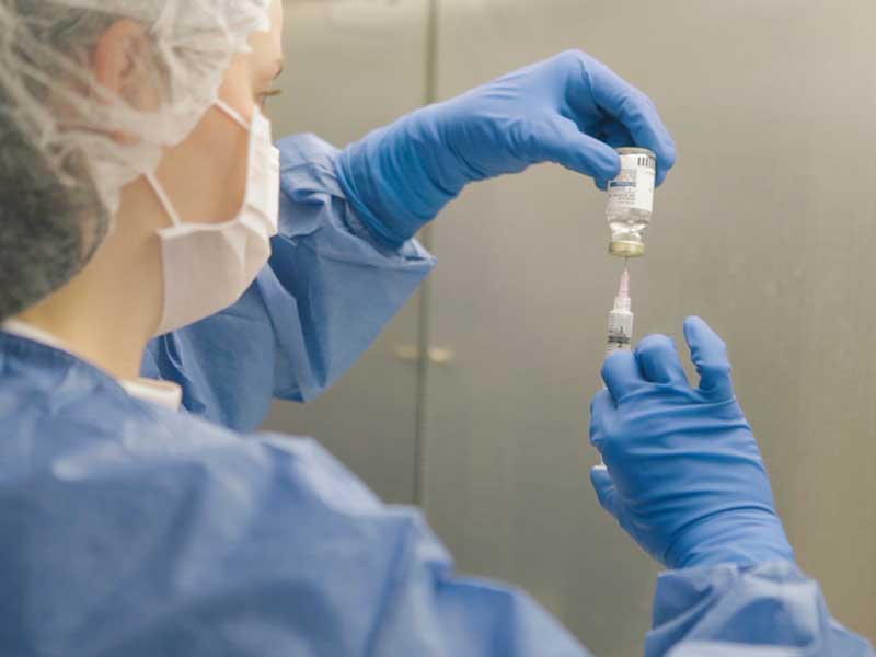 a lab worker in sterile dress extracts liquid from a bottle using a syringe