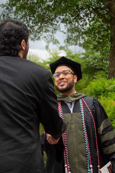 A graduate in cap and gown smiles while shaking hands with another person at an outdoor ceremony