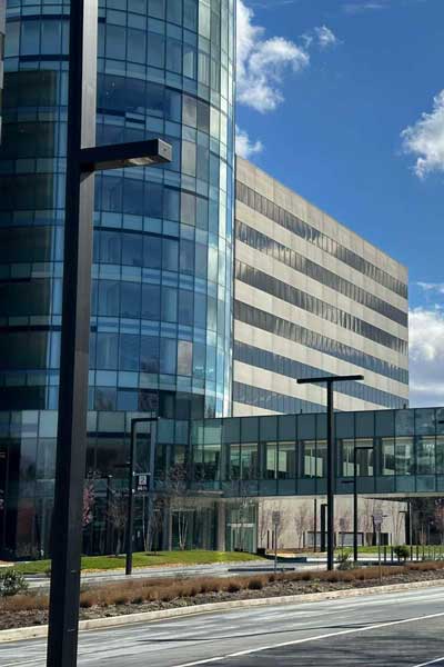 Modern multistory building with a curved glass facade, adjoining rectangular structure and enclosed walkway beside a roadway