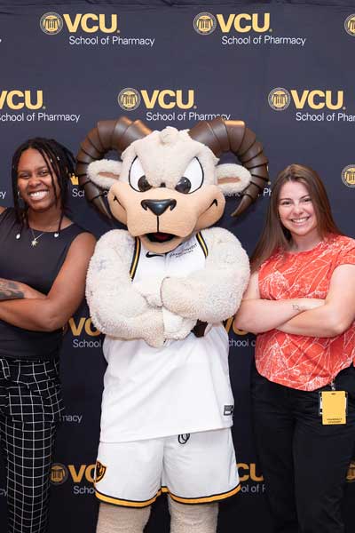 V.C.U. mascot Rodney the Ram poses with a group of two people standing with arms crossed in front of a V.C.U. School of Pharmacy backdrop